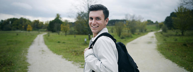 Ein junger Mann mit Rucksack steht an einem sich gabelnden Feldweg, der von Wiese und Bäumen umsäumt ist. Der junge Mann blickt seitlich zurück und lächelt in die Kamera.