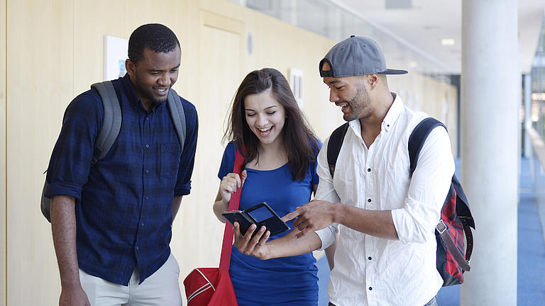 Eine junge Frau und zwei junge Männer stehen in einem Flur zusammen und schauen gemeinsam auf ein Smartphone, 
