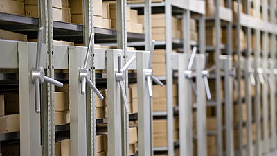 Seitlicher Blick in einen Gang eines Archivs, auf der linken Seite stehen mehrere verschiebbare Regale, die mit braunen Archivkartons bestückt sind.