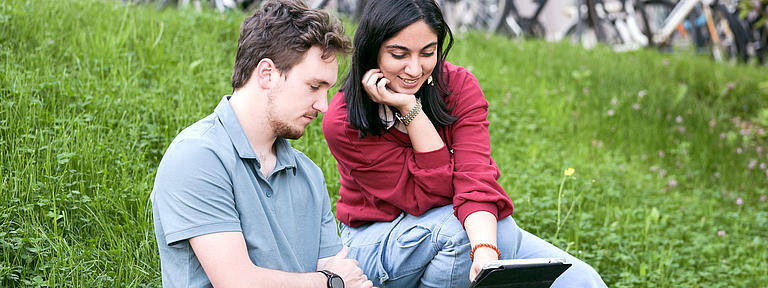 Eine Studentin und ein Student sitzen im grünen Gras mit Tablet.