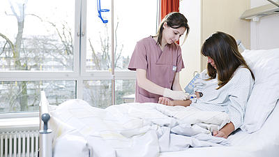 Eine junge Frau mit braunen Haaren sitzt in einem Krankenhausbett in einem lichtdurchfluteten Krankenzimmer vor einem groÃŸen Fenster. Neben ihr steht eine junge braunhaarige Krankenschwester.
