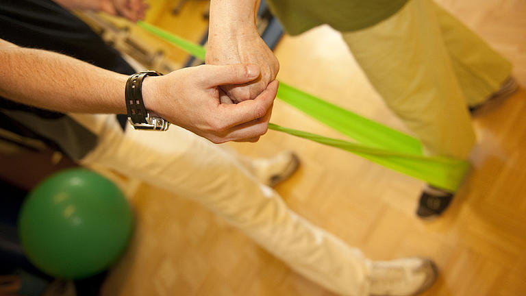 Auf hellem Holzboden sieht man zwei männlich Personen brustabwärts. Die eine Person wird von der anderen bei einer Übung mit einem leuchtend grünen Fitnessband angeleitet