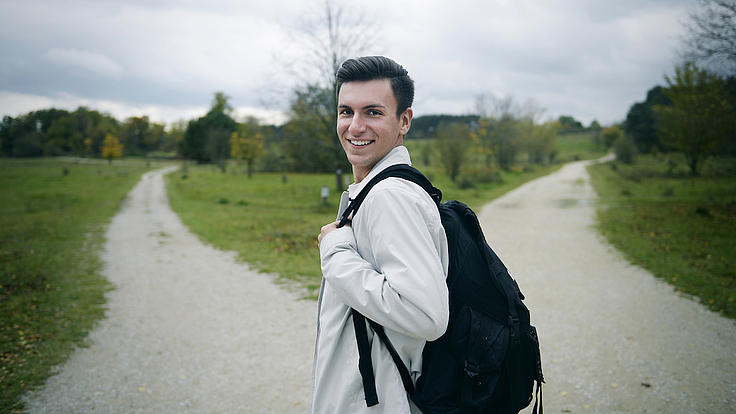 Ein junger Mann mit Rucksack steht an einem sich gabelnden Feldweg, der von Wiese und Bäumen umsäumt ist. Der junge Mann blickt seitlich zurück und lächelt in die Kamera.