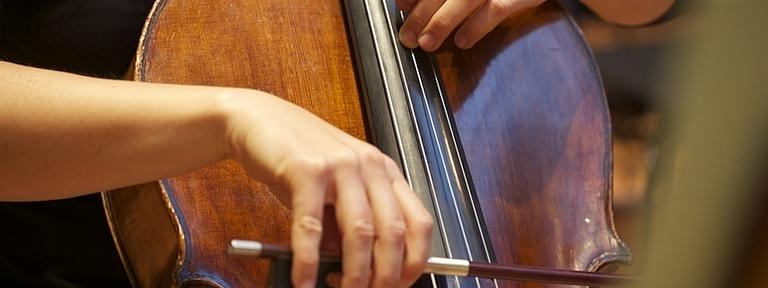 Nahaufnahme eines Cellos, das von einem Mann gehalten wird. Eine Hand hält einen Bogen über das Instrument. Die andere hält die Saiten fest.
