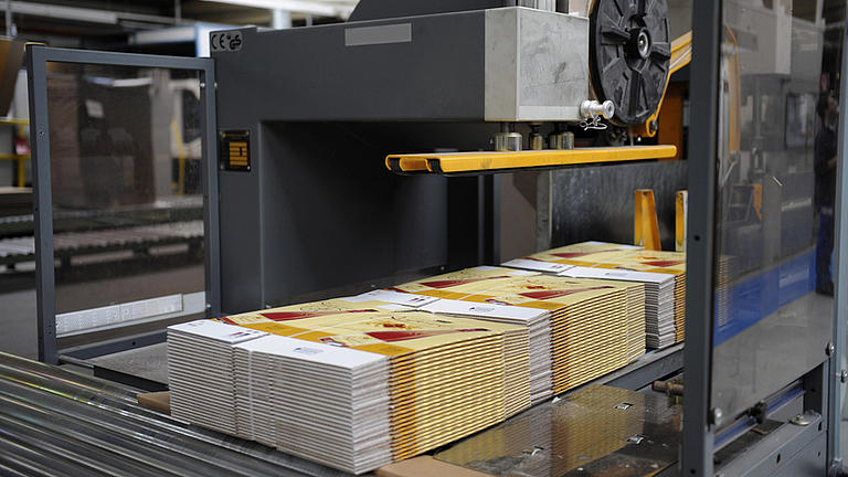 Auf einem Förderband unter einer dunklen Maschine für die Herstellung von Kartonagen liegen drei Stapel mit fertigen Kartons. Die Stapel werden von Kunststoffbändern zusammengehalten.