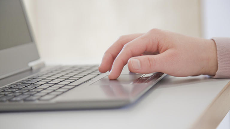 Eine männliche Hand liegt auf einen silbergrauen Laptop, der auf einem weißen Schreibtisch steh