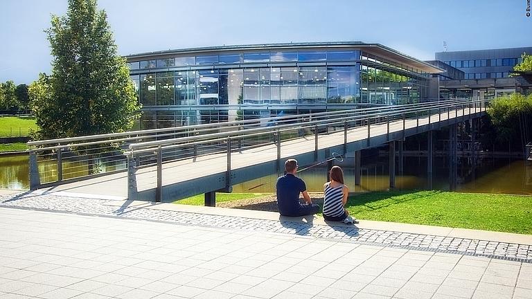 Eine Studentin und ein Student sitzen vor einem gläsernen Hochschulgebäude.