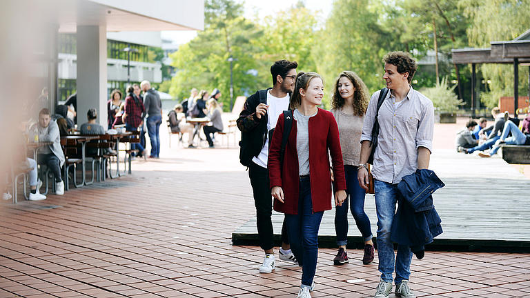 Zwei junge Frauen und zwei junge Männer laufen auf einem breiten Weg. Auf der linken Seite stehen Tische im Freien, an denen mehrere Personen sitzen