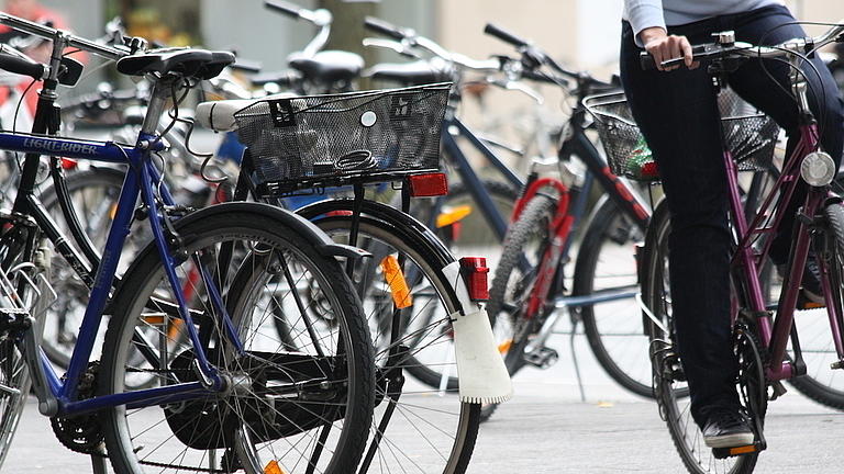 Verschiedene abgestellte Fahrräder auf einem Platz, auf der rechten Bildseite erkennt man die Beine einer vorbeifahrenden Radfahrerin.