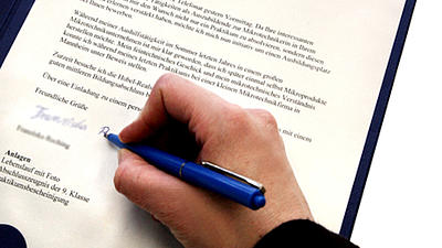 Nahaufnahme eines Bewerbungsanschreibens in einer Mappe, eine Hand hält einen blauen Kugelschreiber und unterschreibt gerade das ausgedruckte Schriftstück.