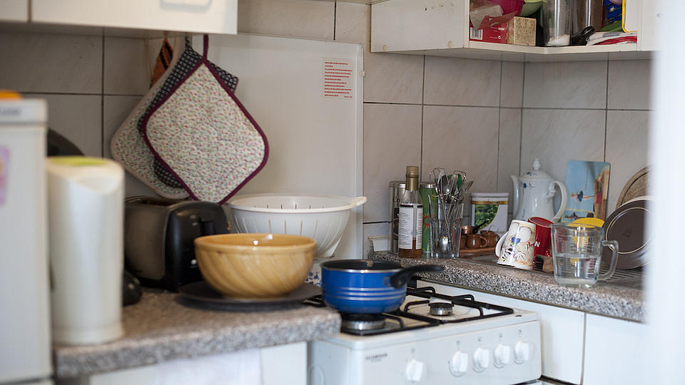 Blick in eine Miniküche mit verschiedenen weißen hängenden und stehenden Küchenschränken sowie einem Gasherd. 