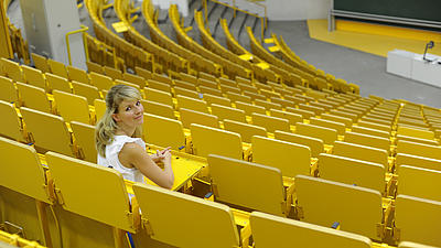 Eine junge Frau sitzt in einem Vorlesungssaal mit gelben Stühlen.