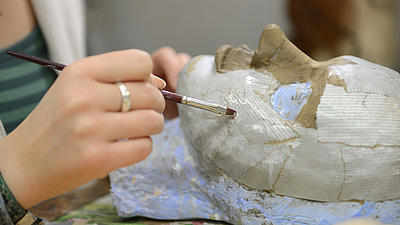 Eine Frauenhand mit Ring am Mittelfinger hält einen Pinsel an eine Maske aus Pappmache, die vor ihr liegt.