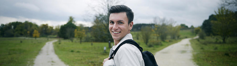 Ein junger Mann mit Rucksack steht an einem sich gabelnden Feldweg, der von Wiese und Bäumen umsäumt ist. Der junge Mann blickt seitlich zurück und lächelt in die Kamera.