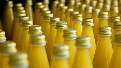 Detailaufnahme von frisch befüllten Orangensaftflaschen in der Produktion. Man blickt über die Glasflaschen mit Metalldeckeln.