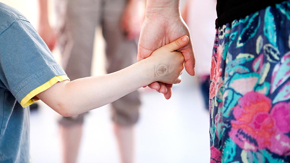 Hand eines Kindes in der Hand einer Frau mit geblümtem Rock. Im Hintergrund eine Person mit kurzer Hose, die die beiden empfängt.