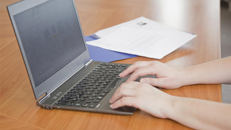 Ein Laptop steht aufgeklappt auf einem Holztisch, die HÃ¤nde einer Person arbeiten auf dem Touchpad.