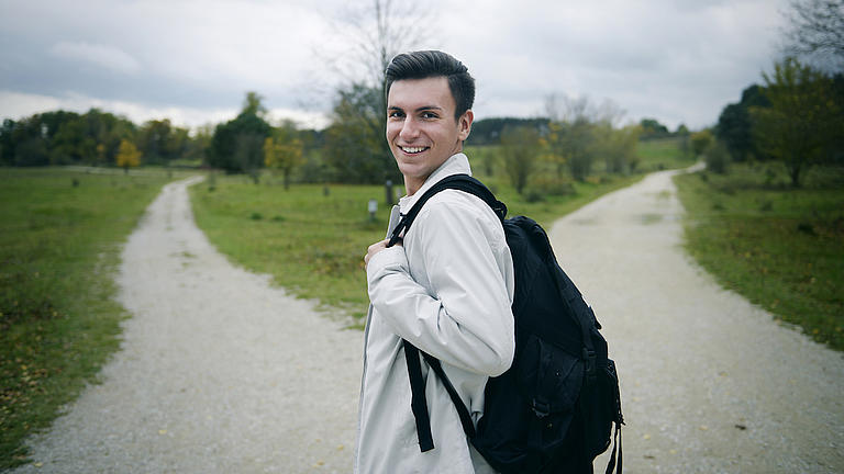 Ein junger Mann mit Rucksack steht an einem sich gabelnden Feldweg, der von Wiese und Bäumen umsäumt ist. Der junge Mann blickt seitlich zurück und lächelt in die Kamera.