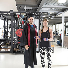 Zwei junge Menschen stehen in einem Fitnessstudio. Die junge Frau trägt Sportklamotten, der junge Mann einen Doktorhut und Robe.