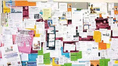 Nahaufnahme einer Pinnwand, die überladen voll ist mit verschiedensten bunten Zetteln, Flyern und Plakaten auf denen Sprachkurse, Vorträge, Partys und andere Aktivitäten angeboten werden.
