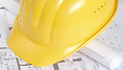 Auf einem aufgefalteten ausgebreiteten Bauplan liegt eine zusammengerollte Papierrolle, auf dieser liegt ein gelber Baustellenhelm.