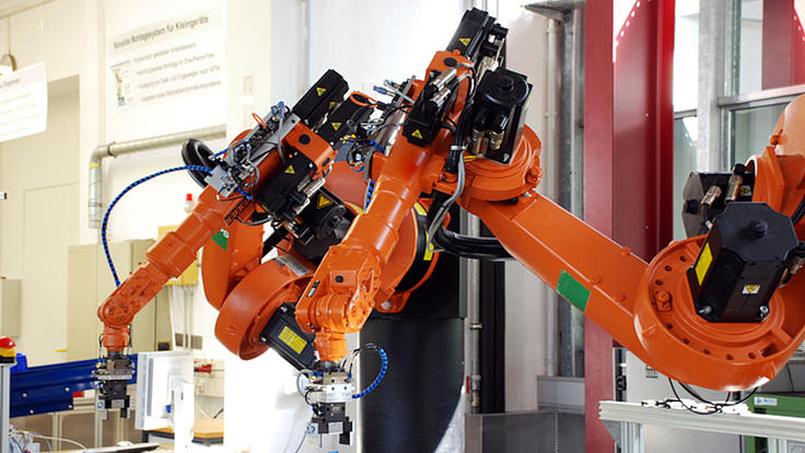 Ein oranger Roboterarm steht  in einer Werkhalle über einem Produktionstisch, auf dem verschiedene Metallteile liegen.