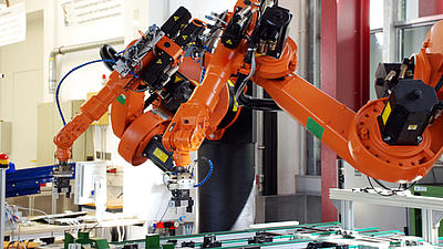 Ein oranger Roboterarm steht  in einer Werkhalle über einem Produktionstisch, auf dem verschiedene Metallteile liegen.
