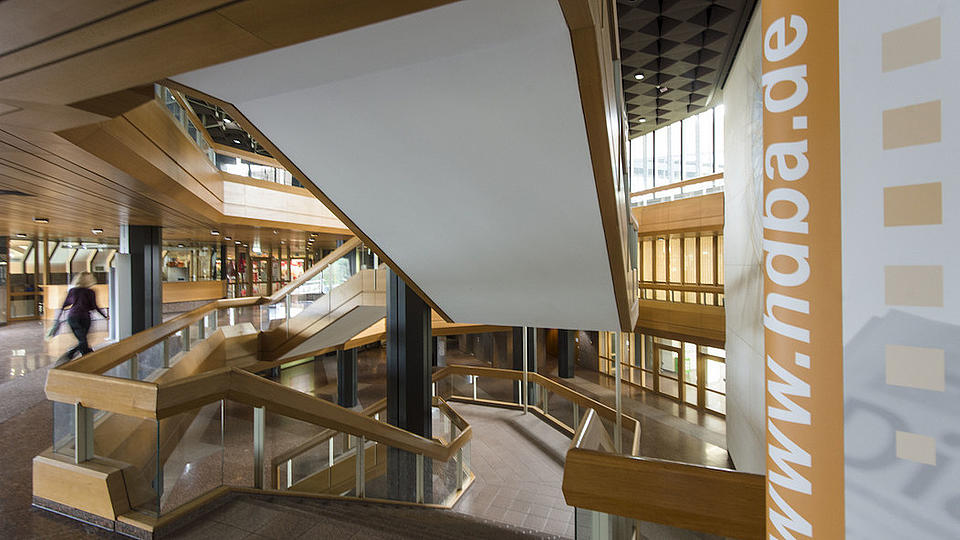 Blick von einem Treppenabsatz auf eine Treppe, die sich in einem großen Gebäude befindet. Das Geländer ist aus Holz gefertigt.
