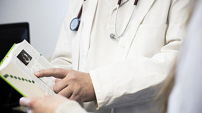 Ein Mann im weißen Kittel, mit Stethoskop um den Hals, zeigt einer anderen Person im Fachbuch Pschyrembel eine Textpassage.