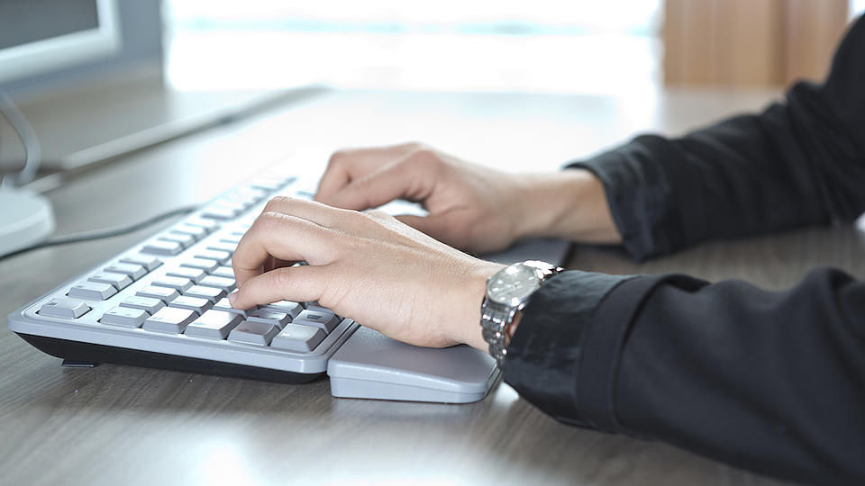 Zwei Hände einer Person schreiben auf einer silbernen Tastatur, die auf einem Tisch vor einem Monitor steht.