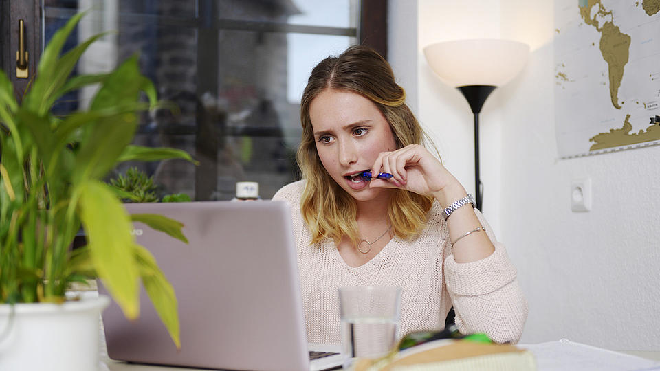 Eine junge Frau sitzt an einem Tisch, auf dem ein geöffneter Laptop steht und schaut auf diesen.