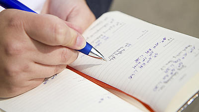Nahaufnahme eines Notizbuches, das von einer Hand gehalten wird. In der andren Hand wird eine blauer Kugelschreiber gehalten.