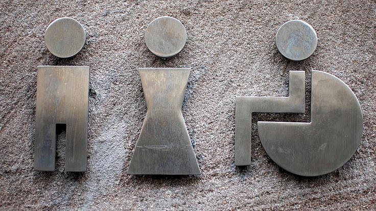 Nahaufnahme dreier Piktogramme aus Metall, auf Stein befestigt. Links ist ein männliches Icon zu sehen, in der Mitte ein weibliches und rechts ein Rollstuhlfahrer.