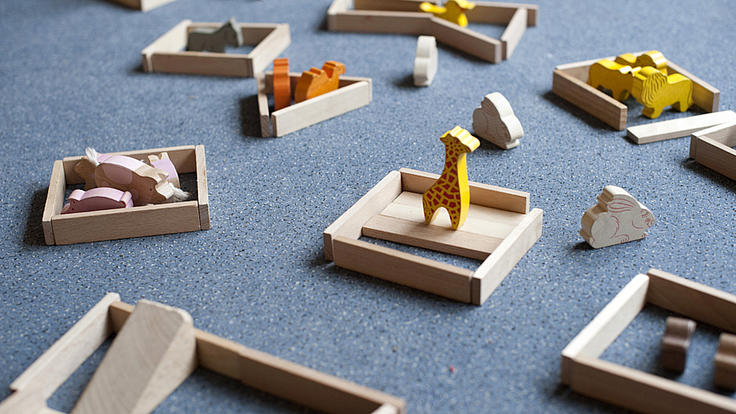 Auf einem grauen Fußboden sind verschiedene Holzspielzeugtiere und andere Holzspielzeuge aufgebaut
