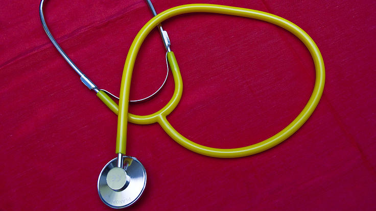 Auf einem roten Tuch liegt ein Stethoskop mit hellgrünen Schlauch und Schlauchgabel, das Bruststück, Ohr- und Federbügel sind metallfarben, die Ohroliven sind gelb.