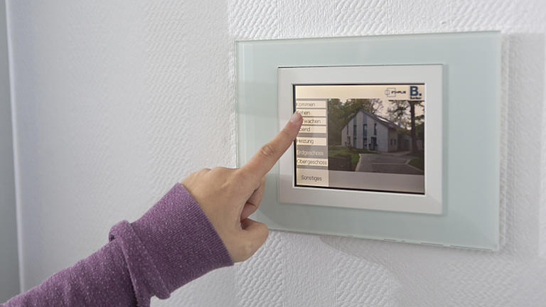 Die Hand einer Frau bedient ein Touchpad einer smarten Steuerungseinheit für ein Einfamilienhaus.