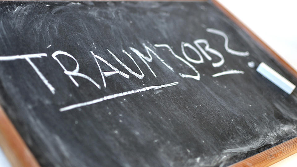 Auf einer schräg liegenden Schreibtafel mit Holzrand steht in Großbuchstaben das Wort "Traumjob"