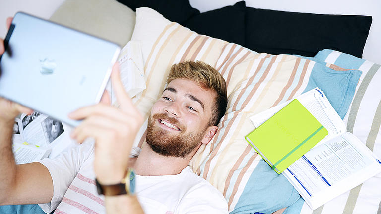 Student liegt mit Tablet und Büchern auf einem Bett 