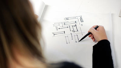 Eine Frau skizziert auf den vor ihr liegenden Zeichenblock mit einem schwarzen Stift Vierecke und Linien.