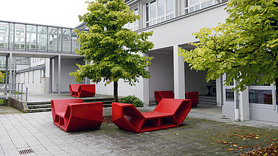 Zu sehen ist ein modernes Hochschulgebäude mit roten Sitzbänken davor.