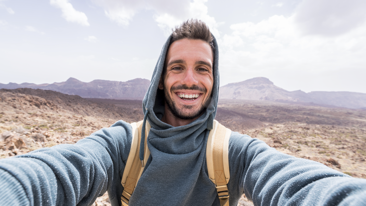 junger Mann lachend mit blauem Kapuzenpulli und Rucksack. Im Hintergrund Natur und Berge