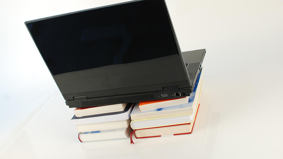 Zwei nebeneinander liegende Buchstapel mit jeweils vier Büchern dienen als Erhöhung für einen aufgeklappten Laptop auf einer weißen Fläche.