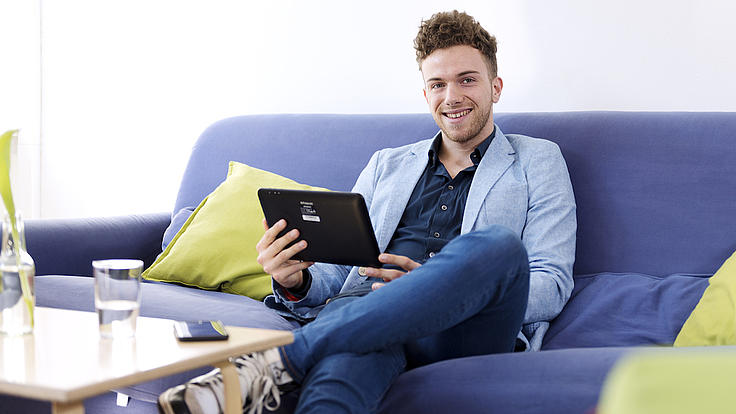 Ein junger Mann sitzt auf einer blauen Couch und hält ein Tablet in seinen Händen. Er hat ein Bein auf das andere gelegt und blickt lächelnd in die Kamera. 
