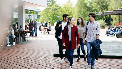 Zwei junge Frauen und zwei junge Männer laufen auf einem breiten Weg vor der Universität.