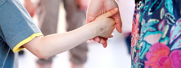 Hand eines Kindes in der Hand einer Frau mit geblümtem Rock. Im Hintergrund eine Person mit kurzer Hose, die die beiden empfängt.