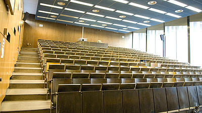 Blick in einen leeren Hörsaal, dessen Sitzreihen aus Holz bestehen. Auch die Wandverkleidung ist holzfarben. An der einen Seite lässt eine riesige Fensterfront Licht herein.