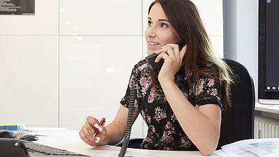 Ein junge Frau sitzt an einem Schreibtisch mit Computer, telefoniert und hat einen Stift in der Hand.