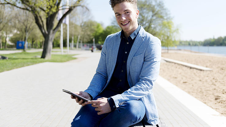 Ein junger Mann sitzt lächelnd auf einer Parkbank im Sonnenschein. In seiner Hand hält er ein Tablet.
