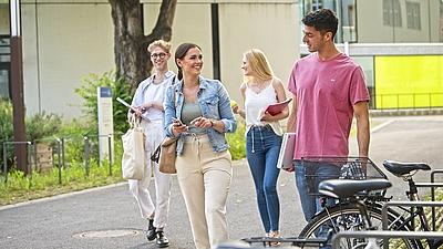 Vier junge Menschen gehen über den Campus vor einem Gebäude und unterhalten sich.