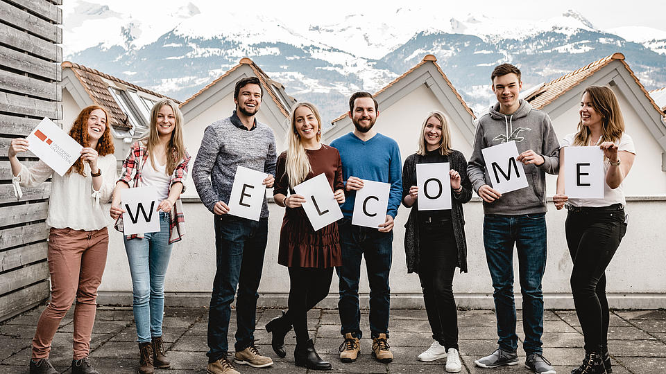Studenten halten ein Werlcome-Schild hoch. Im Hintergrund die Berge mit leichten Schneespitzen.